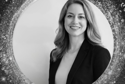 A smiling woman with long hair and wearing a blazer is framed within a circle of star-like points of light.