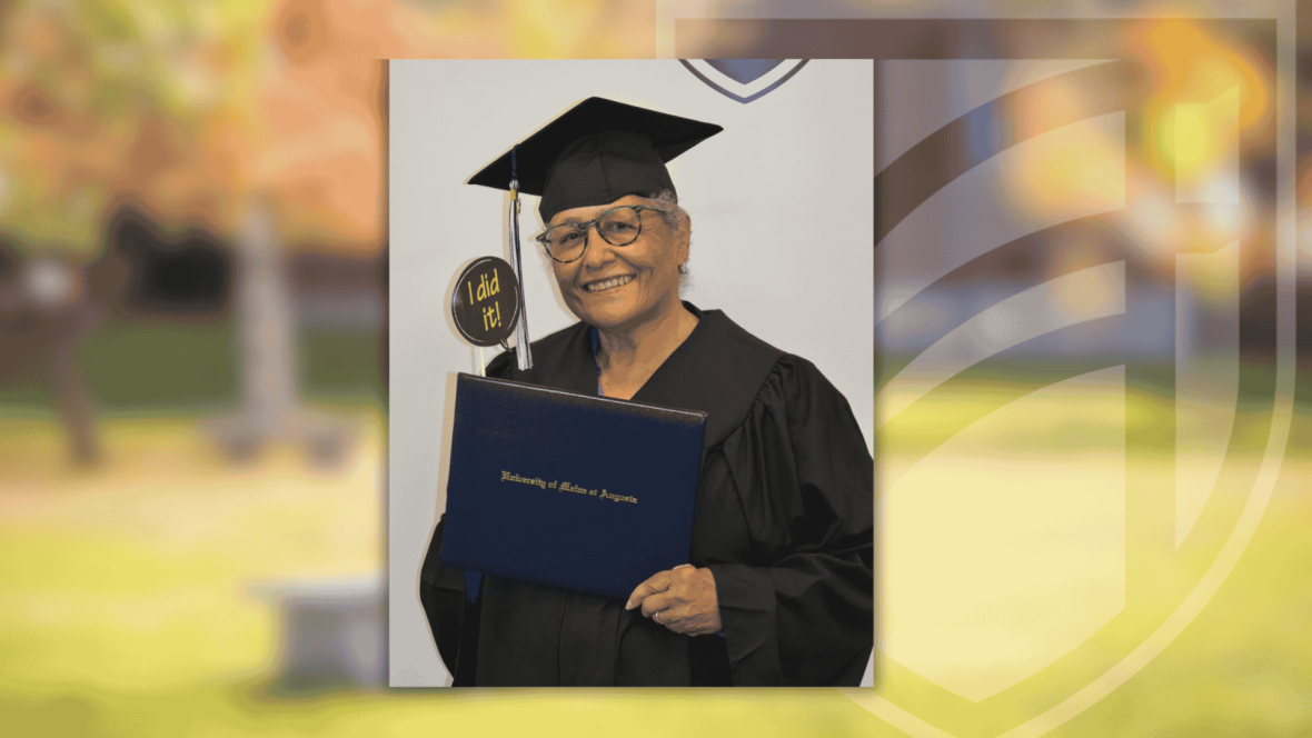 Mimi wearing a graduation cap and gown smiles while holding a University of Maine at Augusta diploma. She also holds a small sign that reads “I did it!”