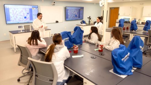 High school students in lab coats listen to a UMA medical laboratory technology demonstration in a clinical laboratory classroom.