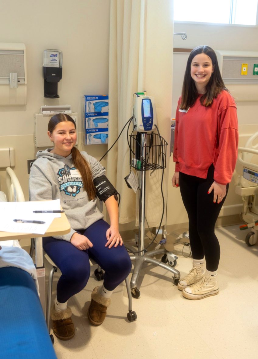 Two high school students in a clinical learning space, with one seated wearing a blood pressure cuff and one standing beside medical equipment.