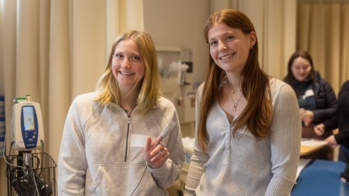 Two smiling high school students stand in a clinical learning space, with one holding a medical device near a vital signs monitor.