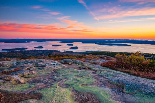 A scenic sunrise over islands in a bay, viewed from a rocky, moss-covered summit with autumn foliage.