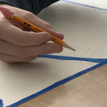 A child's hand is holding a sharpened yellow pencil with a pink eraser, writing on a large sheet of light-colored paper taped off with blue painter’s tape into triangular sections