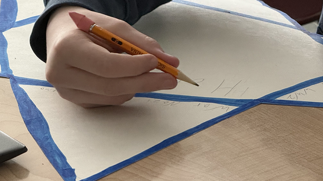 A child's hand is holding a sharpened yellow pencil with a pink eraser, writing on a large sheet of light-colored paper taped off with blue painter’s tape into triangular sections