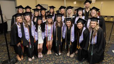 19 dental program graduates wearing caps and gowns with purple sashes and colorful cords pose for a group photo.