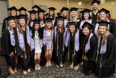 19 dental program graduates wearing caps and gowns with purple sashes and colorful cords pose for a group photo.