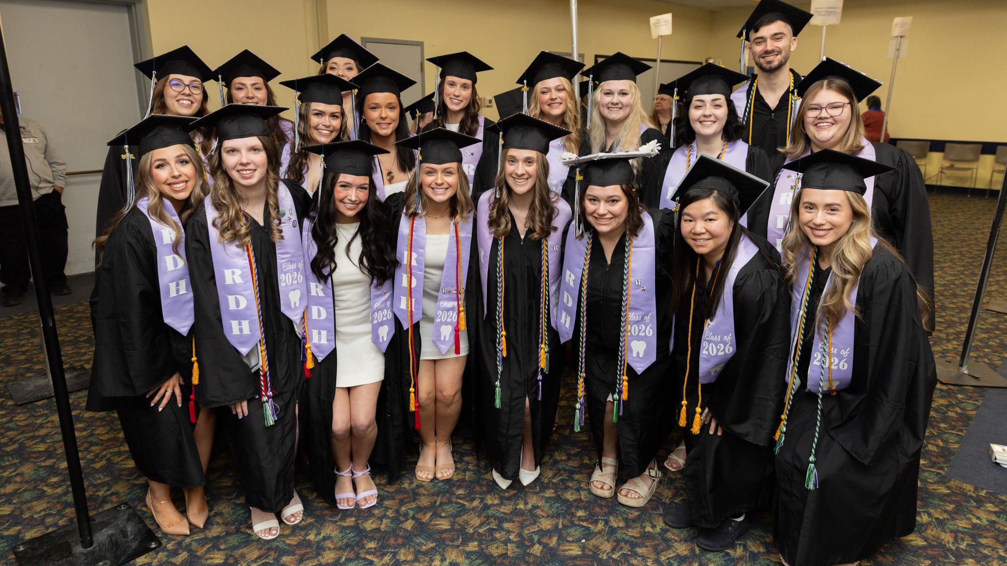 19 dental program graduates wearing caps and gowns with purple sashes and colorful cords pose for a group photo.