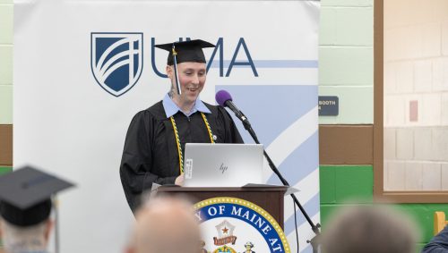 A graduate in a black cap and gown stands at a podium and speaks into a microphone during a UMA commencement ceremony.