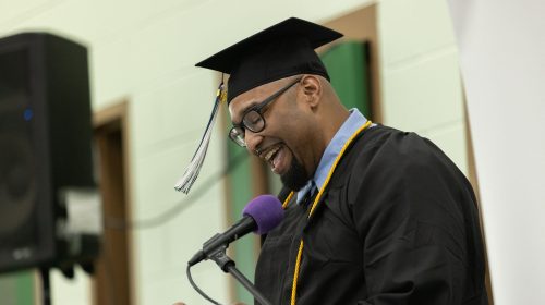 A graduate in a black cap and gown smiles while speaking at a microphone during a commencement ceremony.