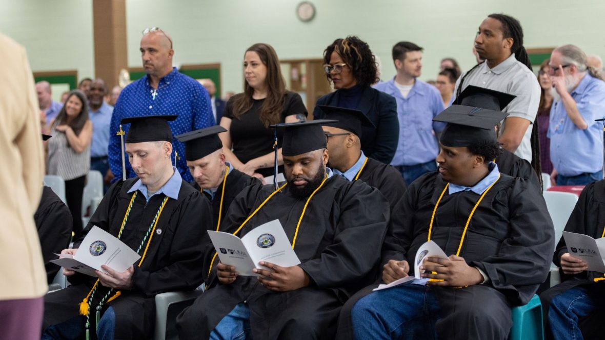 Graduates in black caps and gowns sit with commencement programs while audience members stand behind them at UMA’s 2026 Commencement Ceremony at the Maine State Prison in Warren.