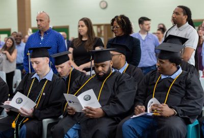 Graduates in black caps and gowns sit with commencement programs while audience members stand behind them at UMA’s 2026 Commencement Ceremony at the Maine State Prison in Warren.
