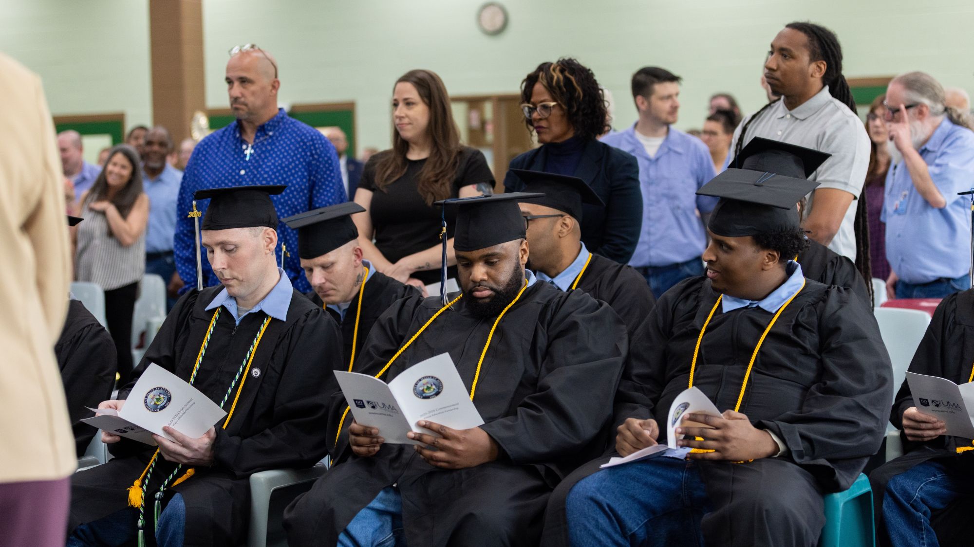Graduates in black caps and gowns sit with commencement programs while audience members stand behind them at UMA’s 2026 Commencement Ceremony at the Maine State Prison in Warren.