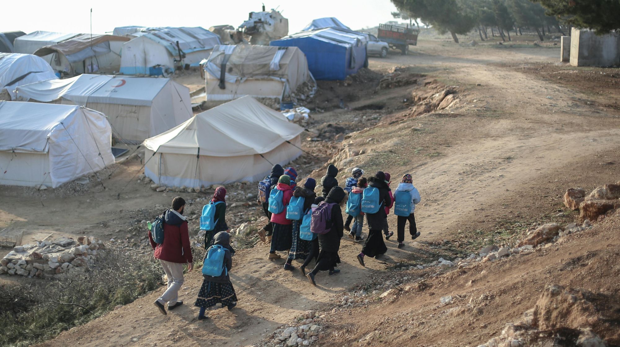 Stock photo of a group of people wearing backpacks, walking up a dirt hill. A tent city is in the background.