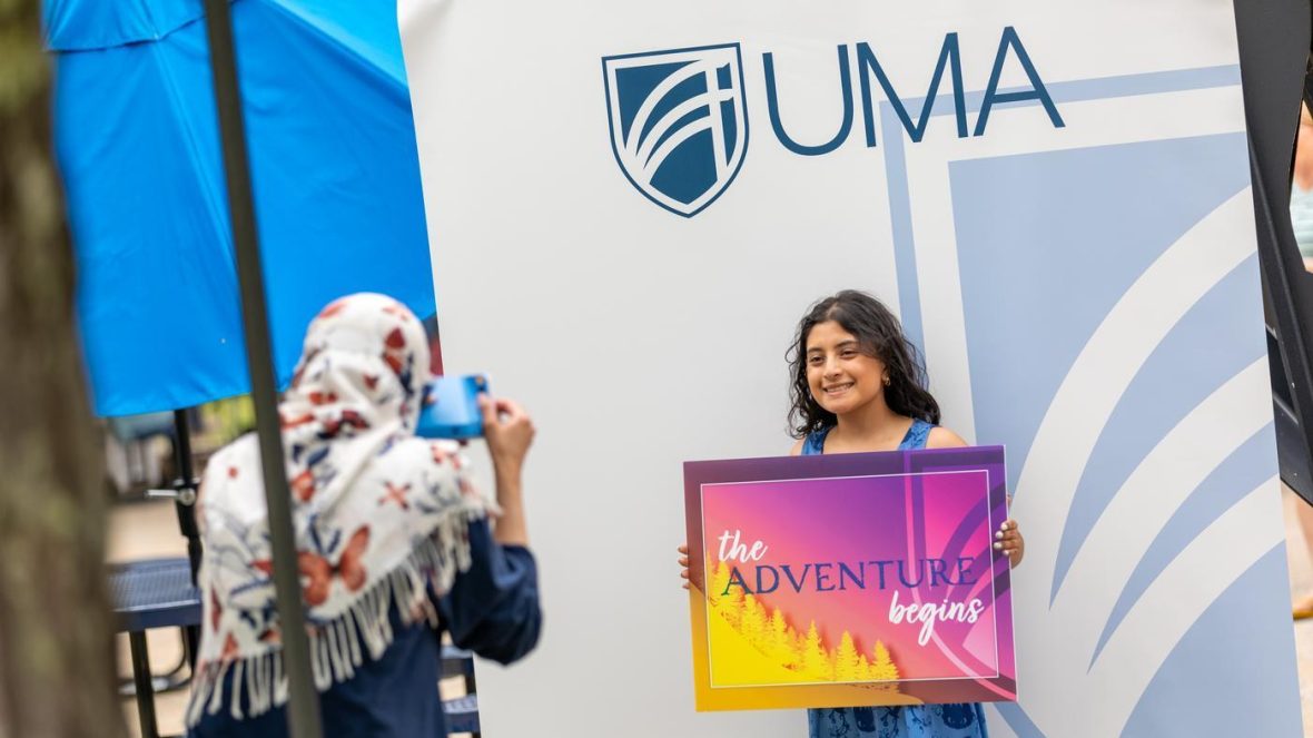 An accepted student posts at the UMA photo booth with a sign that days "The Adventure Begins."