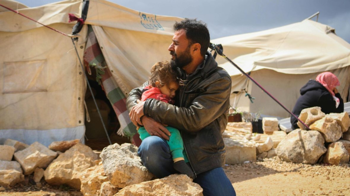A man kneels on a rock while comforting a child in his arms. Refugee tents in the background.