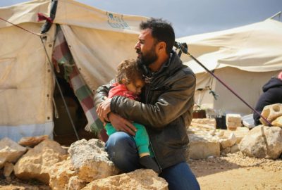 A man kneels on a rock while comforting a child in his arms. Refugee tents in the background.