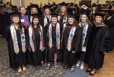 Eight University of Maine at Augusta Bachelor of Science in Veterinary Technology graduates from the Class of 2026 stand together in caps, gowns, honor cords, and stoles before commencement. Two faculty members, one wearing a blue shirt and another in academic regalia, stand with the graduates. The entire graduating class achieved a 100% first-time pass rate on the Veterinary Technician National Examination (VTNE).