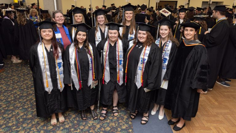 Eight University of Maine at Augusta Bachelor of Science in Veterinary Technology graduates from the Class of 2026 stand together in caps, gowns, honor cords, and stoles before commencement. Two faculty members, one wearing a blue shirt and another in academic regalia, stand with the graduates. The entire graduating class achieved a 100% first-time pass rate on the Veterinary Technician National Examination (VTNE).