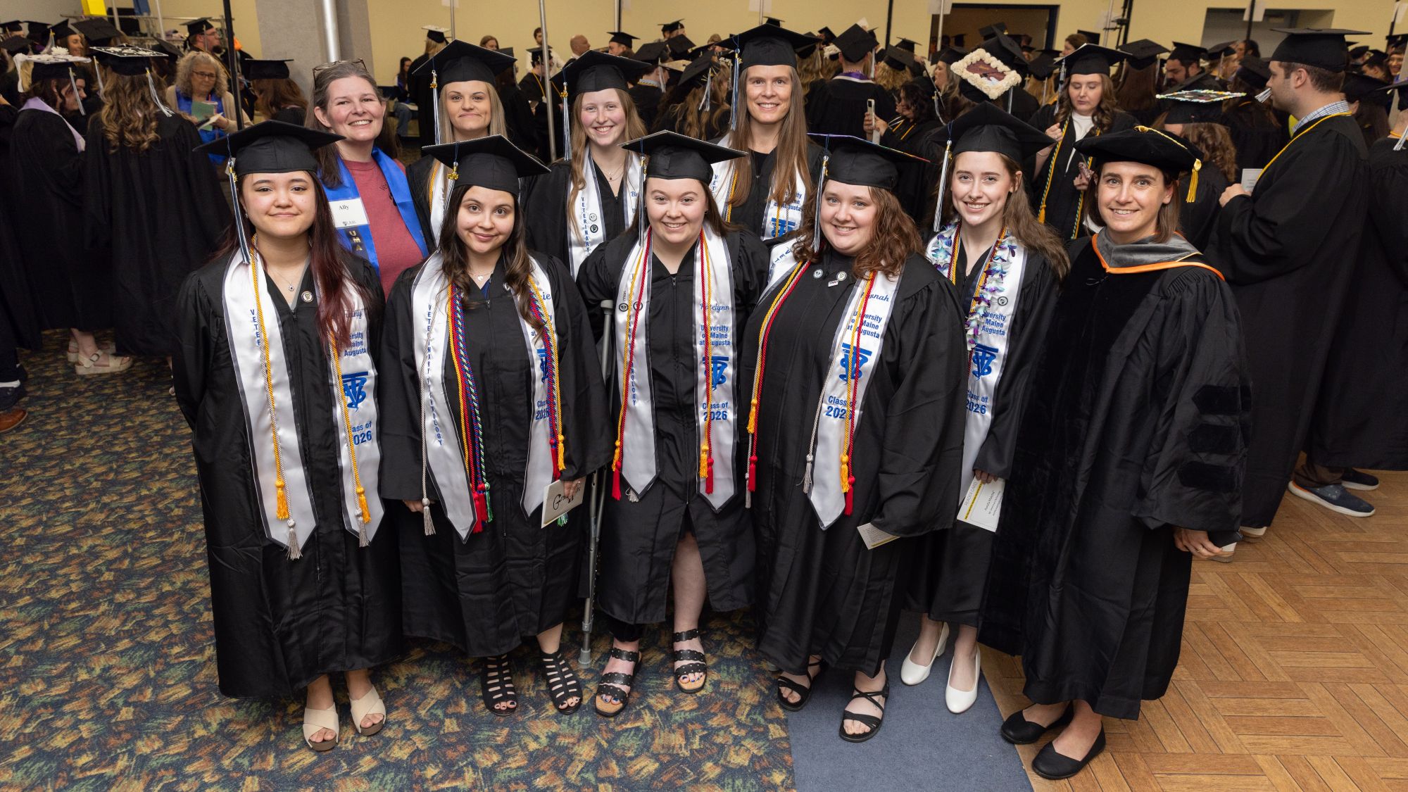 Eight University of Maine at Augusta Bachelor of Science in Veterinary Technology graduates from the Class of 2026 stand together in caps, gowns, honor cords, and stoles before commencement. Two faculty members, one wearing a blue shirt and another in academic regalia, stand with the graduates. The entire graduating class achieved a 100% first-time pass rate on the Veterinary Technician National Examination (VTNE).