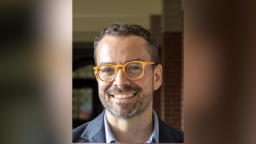 Dan McGloin smiles for a portrait while wearing a dark blazer, light blue shirt, and bright yellow eyeglasses. He is standing in front of a softly blurred campus building.