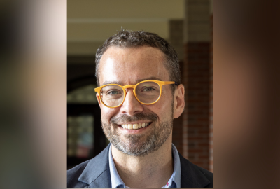 Dan McGloin smiles for a portrait while wearing a dark blazer, light blue shirt, and bright yellow eyeglasses. He is standing in front of a softly blurred campus building.