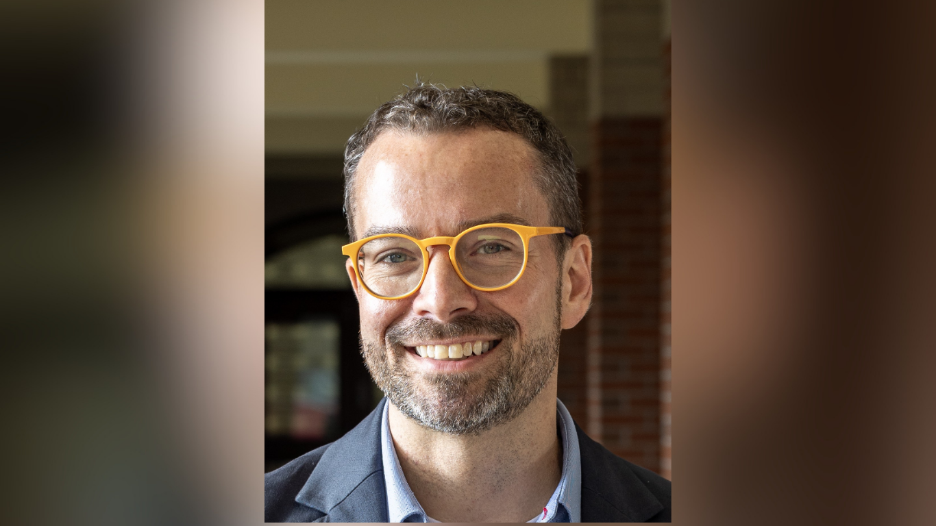 Dan McGloin smiles for a portrait while wearing a dark blazer, light blue shirt, and bright yellow eyeglasses. He is standing in front of a softly blurred campus building.