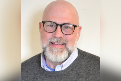 Head-and-shoulders portrait of Robert Bernheim, Ph.D., associate professor of Holocaust and Genocide Studies at the University of Maine at Augusta. He is smiling and wearing black-framed glasses, a gray sweater, and a collared shirt against a light background.