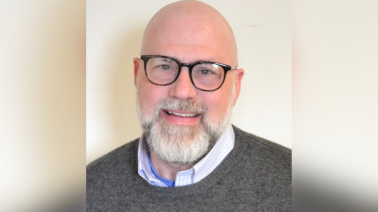 Head-and-shoulders portrait of Robert Bernheim, Ph.D., associate professor of Holocaust and Genocide Studies at the University of Maine at Augusta. He is smiling and wearing black-framed glasses, a gray sweater, and a collared shirt against a light background.