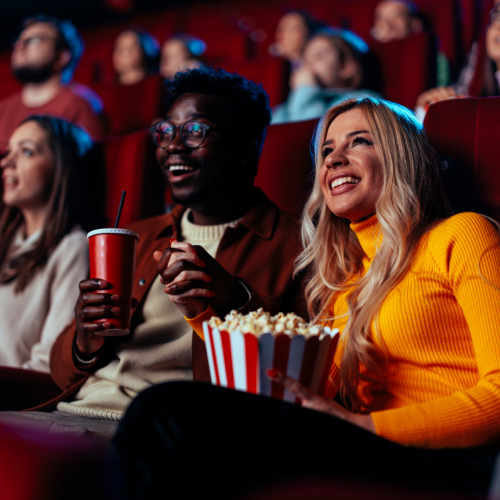 A diverse group of people sits in a movie theater. A couple in the foreground is looking at the screen and sharing popcorn and a red drink.
