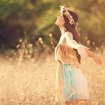 A young woman wearing a floral crown stands with her arms outstretched in a field of dry grass, her face tilted towards the sun.