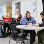 Six people sit and stand around tables in a campus space, talking and reviewing materials together. A rainbow Pride flag hangs on the wall behind them.