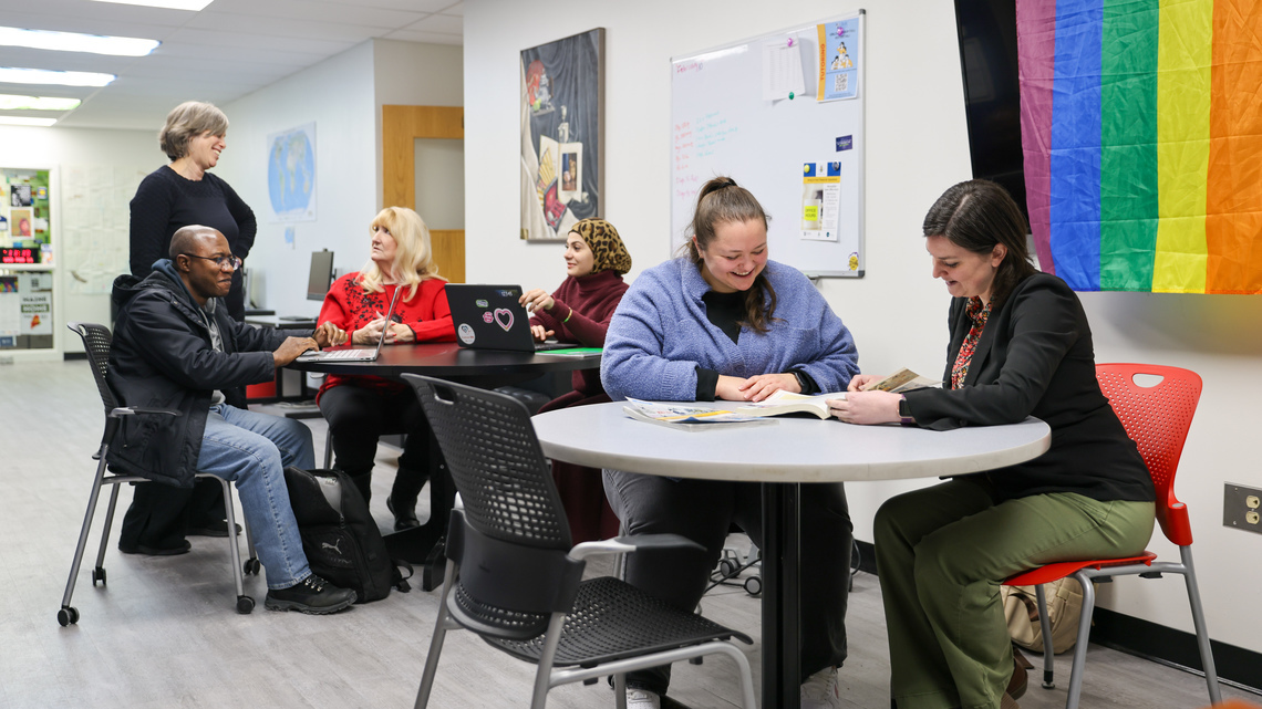 Six people sit and stand around tables in a campus space, talking and reviewing materials together. A rainbow Pride flag hangs on the wall behind them.