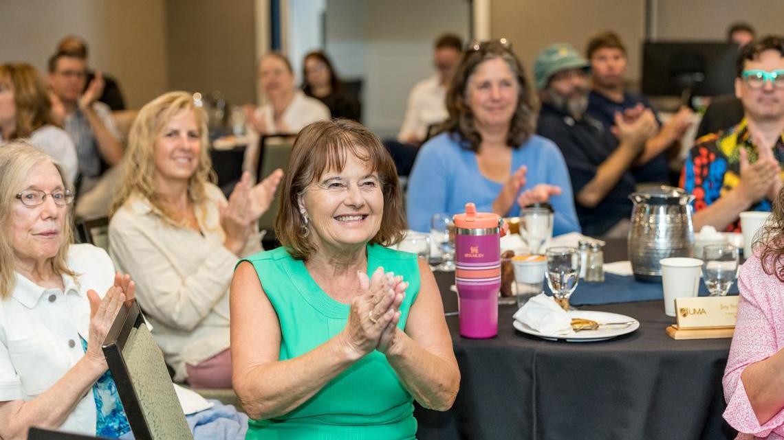 A group of people seated around tables at an indoor gathering applaud. In the foreground, a smiling woman in a bright green sleeveless top claps her hands while others nearby join in.