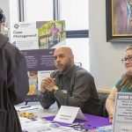 Three people speak at an information table during a job and resource fair. Brochures, hiring information, pens, and other materials are displayed on the table.