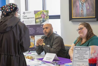 Three people speak at an information table during a job and resource fair. Brochures, hiring information, pens, and other materials are displayed on the table.