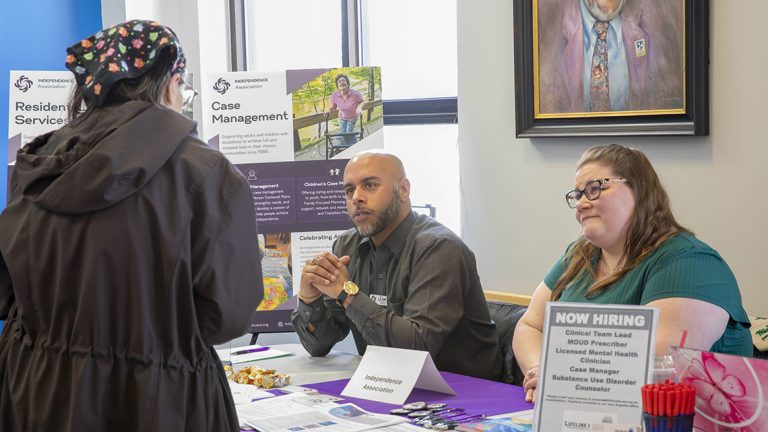 Three people speak at an information table during a job and resource fair. Brochures, hiring information, pens, and other materials are displayed on the table.