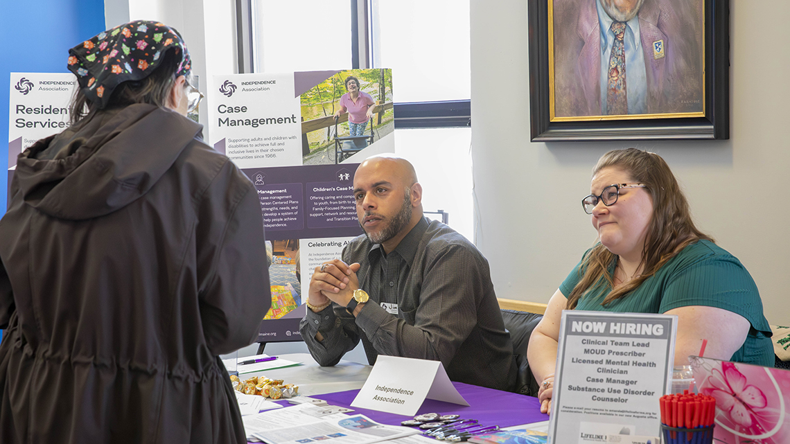Three people speak at an information table during a job and resource fair. Brochures, hiring information, pens, and other materials are displayed on the table.