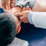 person laying face-down on a medical table, a provider pushes their thumbs along the back of their neck
