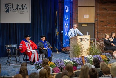 Speaker John Warner stands at a podium during UMA Convocation as faculty in academic regalia sit on stage and an ASL interpreter signs beside him. Students and other attendees watch from the audience.