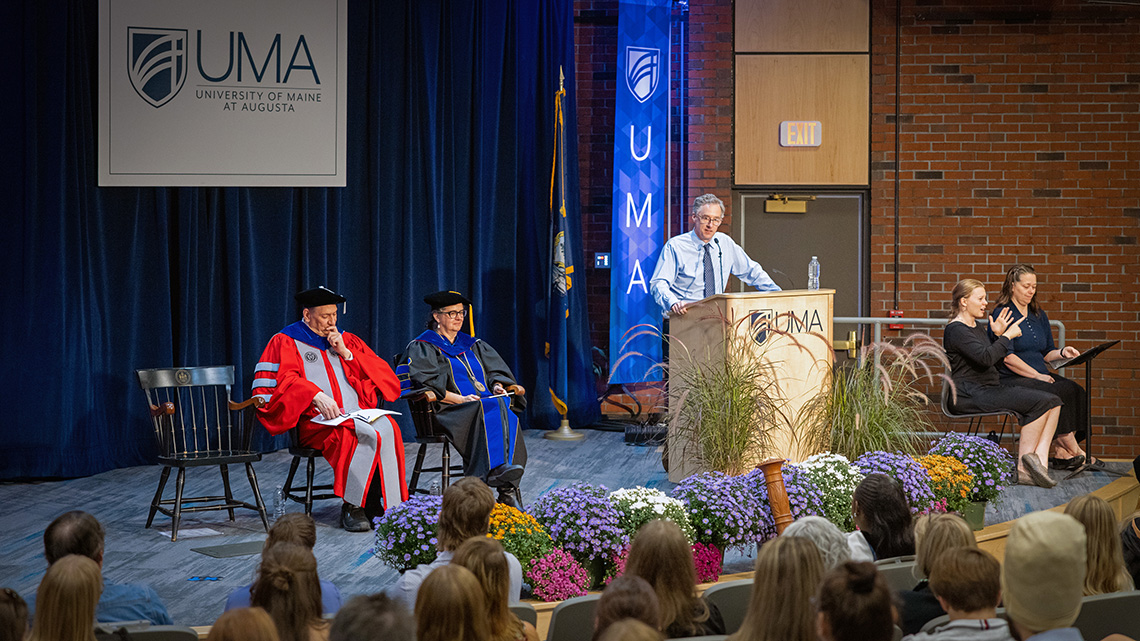 Speaker John Warner stands at a podium during UMA Convocation as faculty in academic regalia sit on stage and an ASL interpreter signs beside him. Students and other attendees watch from the audience.