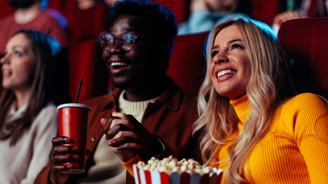 A diverse group of people sits in a movie theater. A couple in the foreground is looking at the screen and sharing popcorn and a red drink.