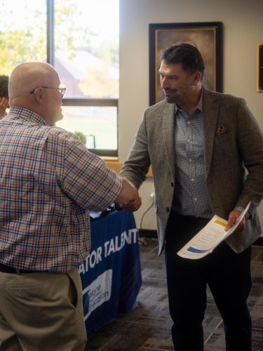 A job seeker in a gray tweed blazer, holding a flyer, shakes hands with a representative at the Maine Department of Education table during UMA’s Fall 2026 Job & Resource Fair.