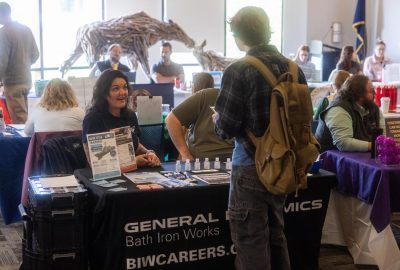 A student with a backpack talks with a smiling recruiter at the General Dynamics Bath Iron Works table, while other job seekers visit employer tables throughout the busy fair.