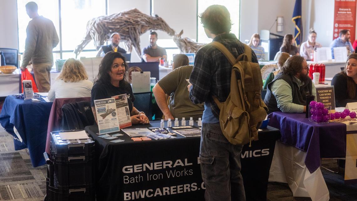A student with a backpack talks with a smiling recruiter at the General Dynamics Bath Iron Works table, while other job seekers visit employer tables throughout the busy fair.