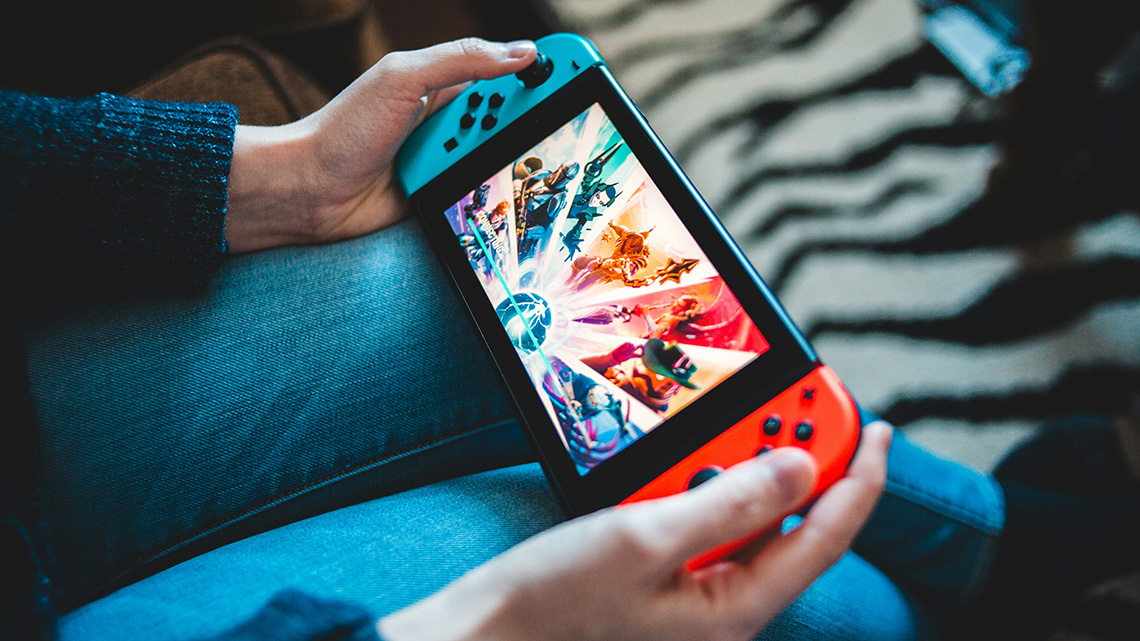 A person seated with a handheld game console resting on their lap, holding it with both hands by the red and blue controllers. A colorful action game with animated characters is on the screen.
