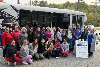 About 20 UMA dental health faculty and students pose outside the UMA Lewiston Center beside a bus and a sign welcoming dental clinic patients, with fall foliage behind them.