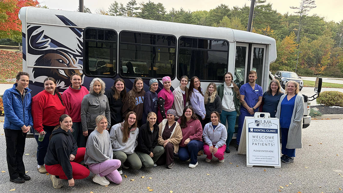 About 20 UMA dental health faculty and students pose outside the UMA Lewiston Center beside a bus and a sign welcoming dental clinic patients, with fall foliage behind them.