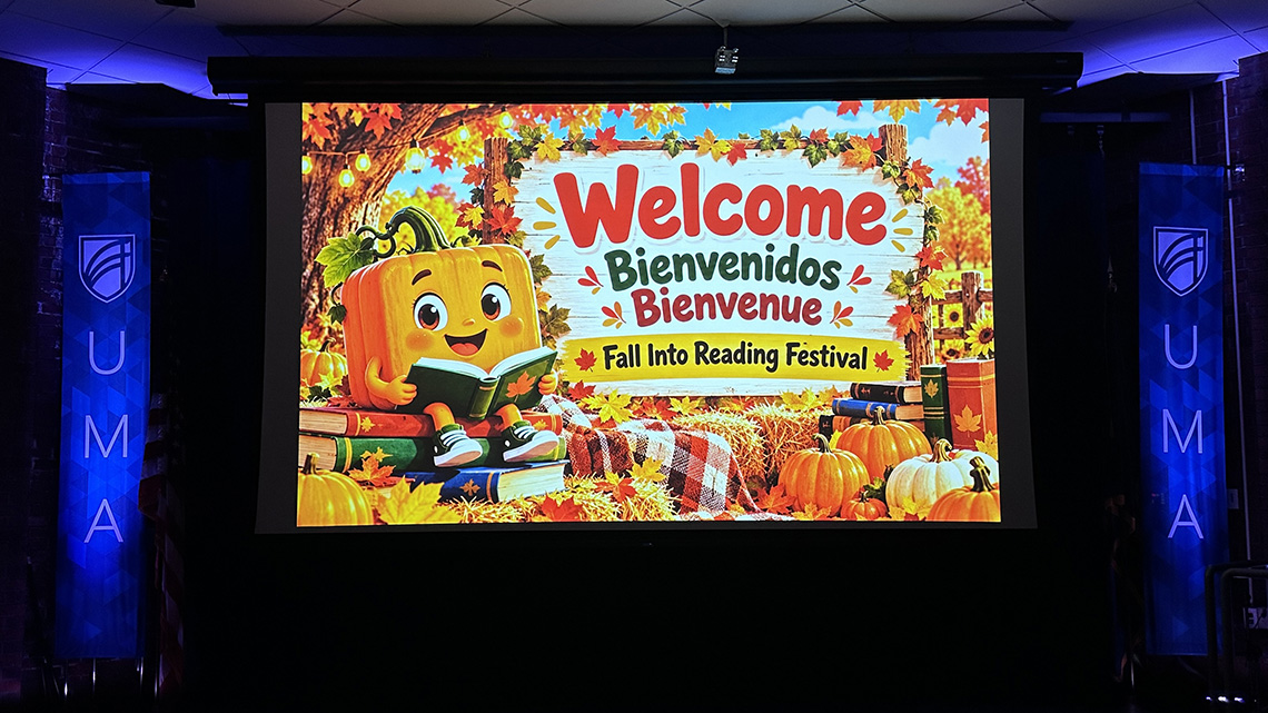 A projection screen displays a colorful autumn-themed slide reading “Welcome, Bienvenidos, Bienvenue: Fall Into Reading Festival,” with a smiling cartoon pumpkin reading a book among fall leaves and pumpkins. UMA banners flank the screen in a darkened event space.
