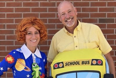 Two people pose in costume against a brick wall. One wears an orange curly wig and a space-themed dress with a toy frog, dressed as Ms. Frizzle from The Magic School Bus; the other wears a yellow school bus costume and yellow shirt as the bus itself. Both are smiling.