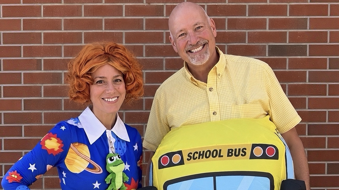 Two people pose in costume against a brick wall. One wears an orange curly wig and a space-themed dress with a toy frog, dressed as Ms. Frizzle from The Magic School Bus; the other wears a yellow school bus costume and yellow shirt as the bus itself. Both are smiling.